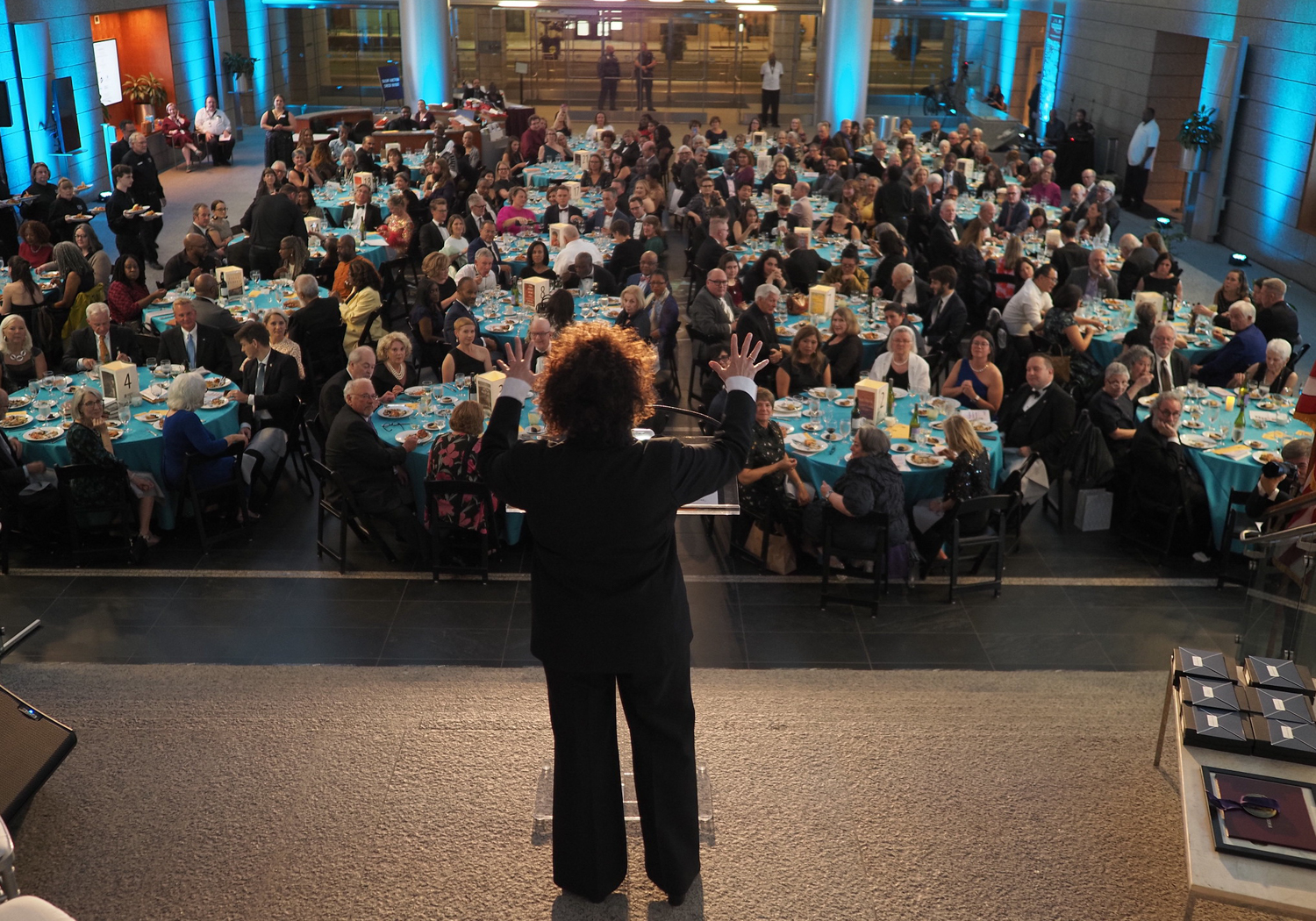 A person stands with their back to the camera, facing a crowd of people sitting at round tables at a large event.