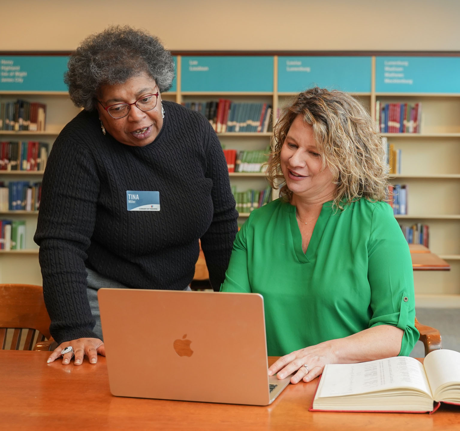 Two women at a table looking at a laptop computer. One woman in a black sweater is standing and the other woman, in a green shirt, is seated.