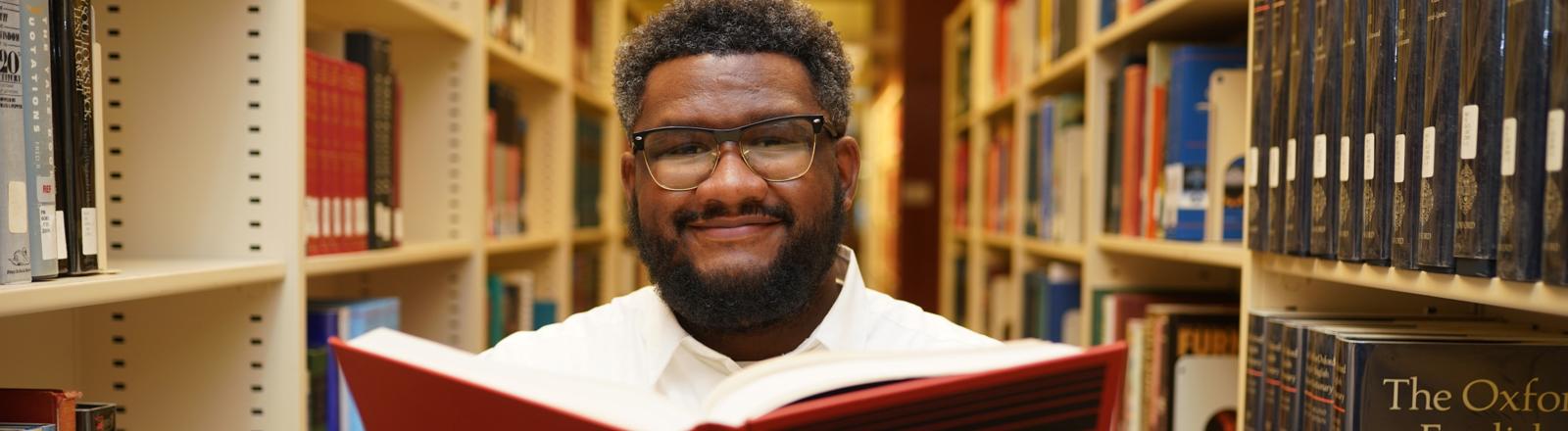 This image shows a man holding a book open while standing in between rows of book shelves.