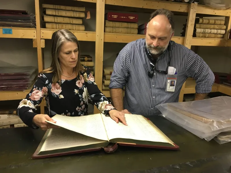 woman and man looking as a bound volume laid open. The woman points to an entry in the volume.